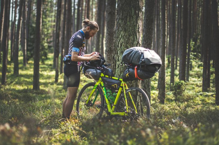 Krunni ja habemega mees kontrollib oma GPS-i männiparkmetsas, täispakitud kollane jalgratas toetub puu peale.
