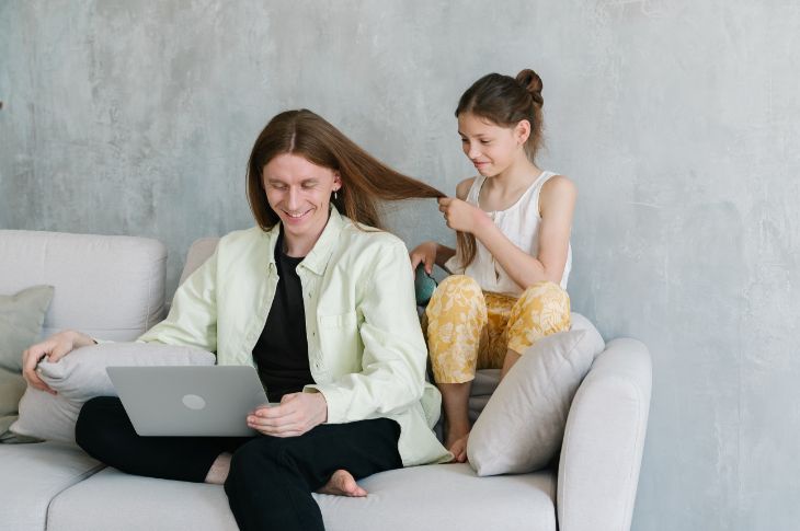 Pikkade juustega isa istub diivanil ja kasutab sülearvutit samal ajal, tütar punub talle patsi.